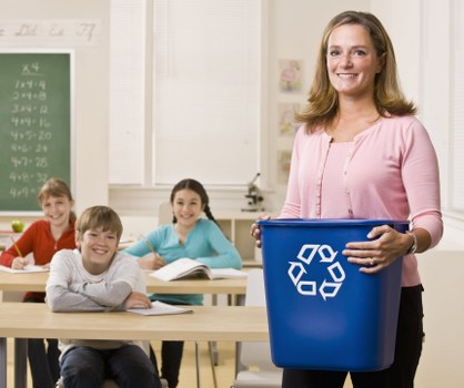 A woman standing in a classroom holding a blue recycling bin with a white recycling symbol, surrounded by three seated children at desks, with a chalkboard and educational posters in the background. The children are smiling and appear engaged, indicating a learning environment. The classroom has natural light coming through windows, and the setting is clean and organized, with the focus on waste segregation and environmental awareness, consistent with rubbish removal themes related to educational or community initiatives in Crouch End.