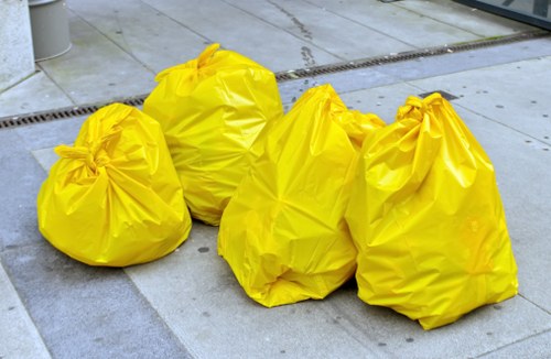 Four large yellow rubbish bags made of durable plastic are stacked on a paved outdoor surface, likely on a driveway or pavement in a residential or commercial area. The bags appear to contain waste materials and are tightly gathered at the top with visible creases and folds. The background shows a concrete surface with a linear drainage grate running across it, suggesting an outdoor environment designed for waste collection. The scene is lit evenly, indicating natural daylight, and the focus is on the bags, which are aligned in a cluster with some leaning against each other. This image is relevant to rubbish removal services, such as those offered by Commercial Waste Crouch End, particularly in the context of managing waste in the Crouch End area, possibly for local businesses or residents needing waste collection. The yellow colour of the bags makes them stand out against the grey paving, emphasizing their purpose related to waste disposal.