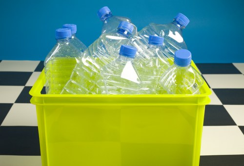 A bright yellow plastic bin filled with several clear plastic bottles that have blue caps, piled unevenly inside. The bottles appear to be partially crushed and are made of transparent PET material, with some showing slight reflections and textural ridges along the sides. The bin is positioned on a black and white checkered floor, with a solid teal or blue background behind it, suggesting an indoor environment such as a recycling or waste collection area in Crouch End. The scene reflects typical rubbish collection imagery associated with waste removal services, highlighting the collection of recyclable plastic bottles. Commercial Waste Crouch End offers services related to rubbish collection, and this image emphasizes the handling of plastic waste materials for recycling or disposal within the local area.