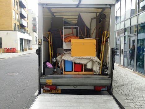 The rear interior of a commercial rubbish removal vehicle parked on a city street shows an open loading space filled with various discarded items. Visible objects include a yellow plastic storage container positioned at the back left, a white fluffy rug draped over some items, and several pieces of furniture such as a black chair with a curved armrest and a wooden or composite table or desk. The items appear to be a mix of household and office waste, with some smaller objects like a black bag and plastic containers also present. The vehicle's interior is lined with a plain, light-colored surface, and the exterior features a yellow and black license plate reading 'KUBLFV.' The surrounding environment includes modern buildings with glass windows and a paved street with a narrow sidewalk to the right, indicating an urban area in London, possibly near Crouch End. This image exemplifies the type of waste collection services offered by Commercial Waste Crouch End for clearing domestic or commercial rubbish in the local area.