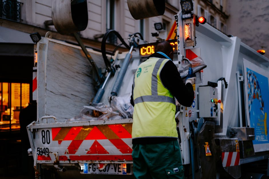 A waste collection worker dressed in a high-visibility yellow vest with reflective strips, black and green protective clothing, and gloves is operating the controls of a large white refuse collection 