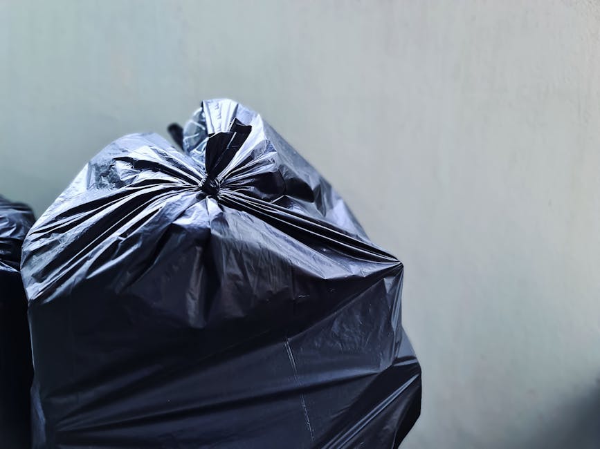 Two large black plastic rubbish bags, filled and tied at the top, are placed on the edge of a pavement in front of a dark wooden fence and a dense green bush. The bags are somewhat crumpled, with visi